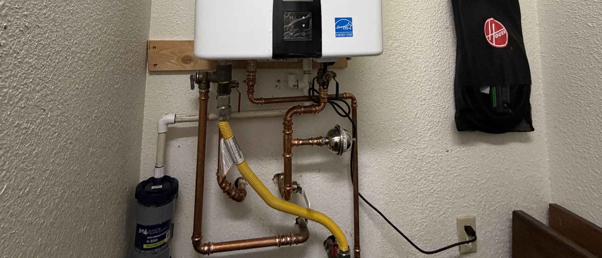 The copper manifold under a tankless water heater: isolation and service valves for flushing scale, a yellow gas flex connector, and an inline filter cartridge on the cold supply