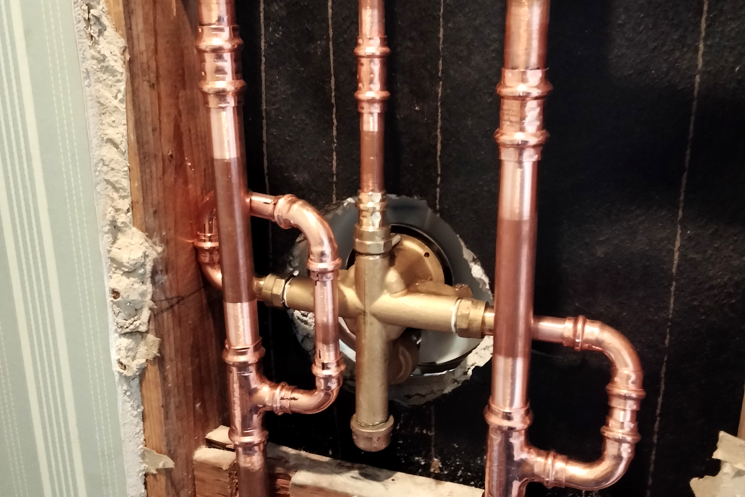 A brass pressure-regulating valve set into new copper supply lines in an opened wall bay, with the drywall cut back to the studs